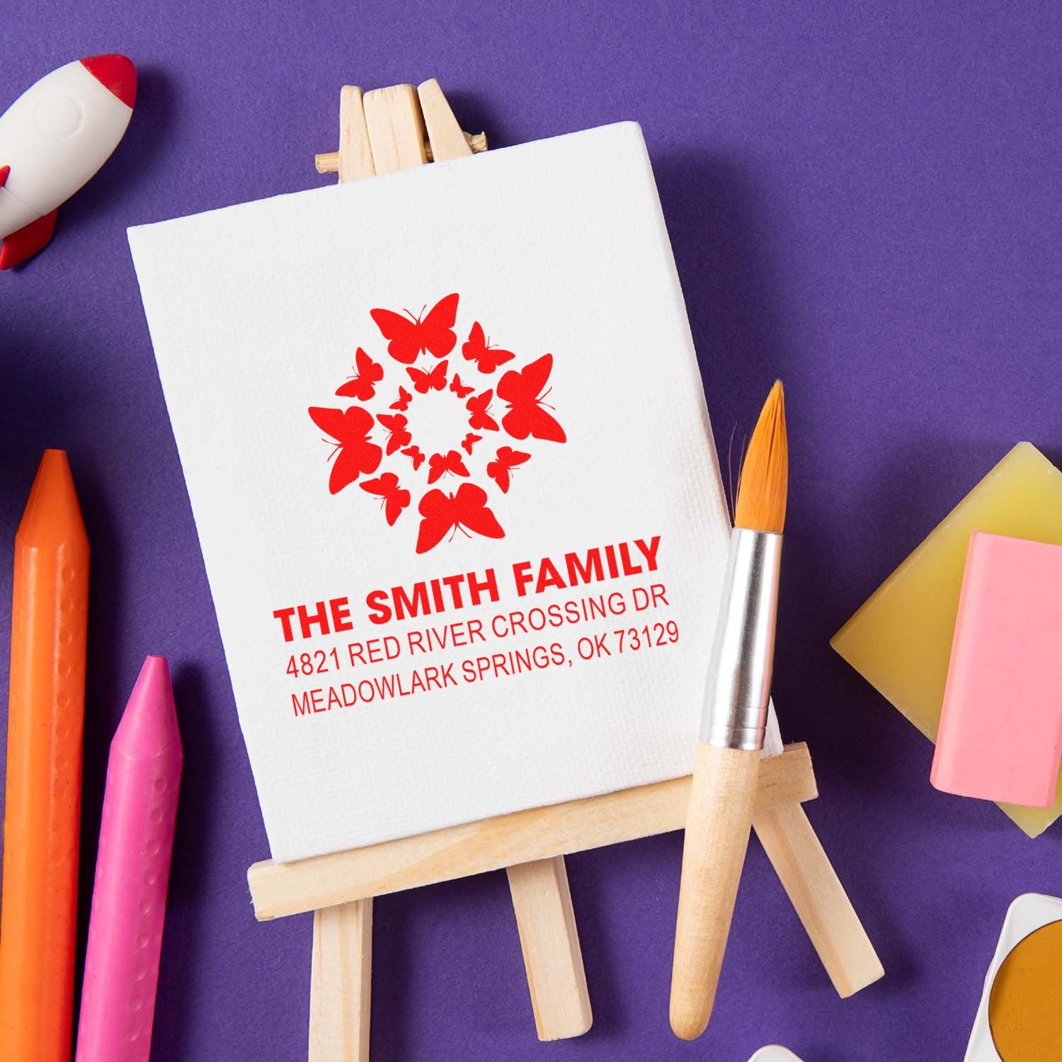 Custom butterfly family address rubber stamp featuring The Smith Family and Meadowlark Springs, displayed on a canvas easel with art supplies.