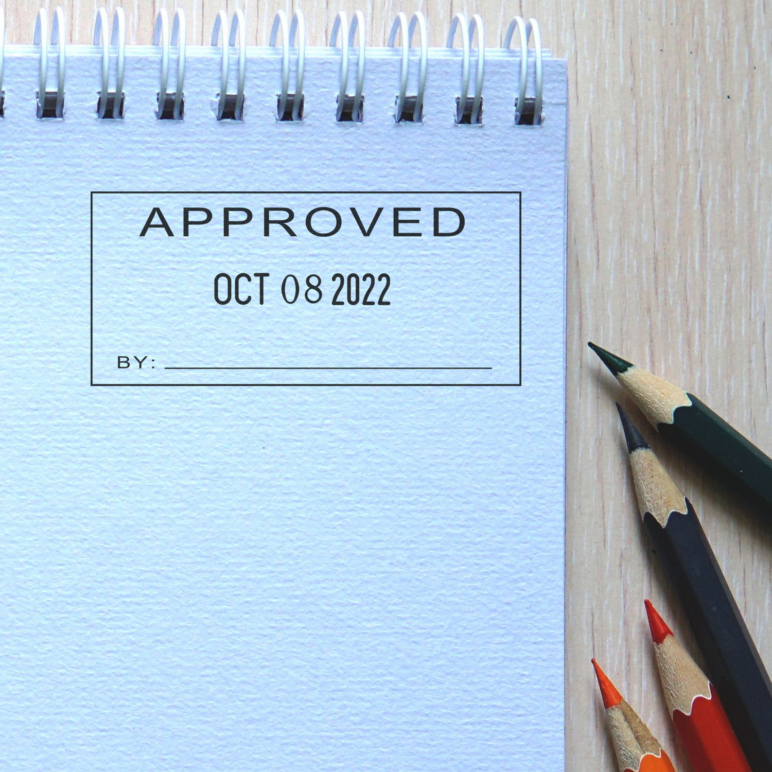 Notebook page with approved stamp and date beside colored pencils on a wooden desk.