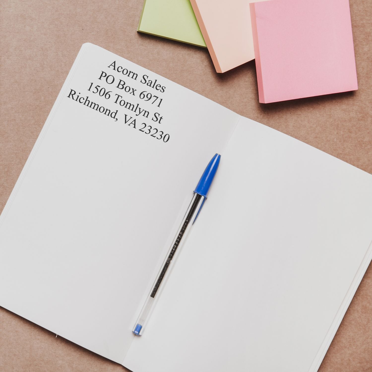 Open notebook with blue pen, printed mailing address, and pastel sticky notes on a brown paper background.