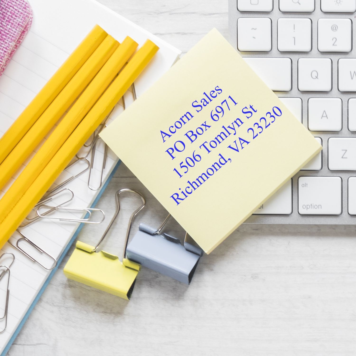 Office desk with keyboard, yellow pencils, paper clips, binder clips, and a sticky note with mailing address details for business correspondence.