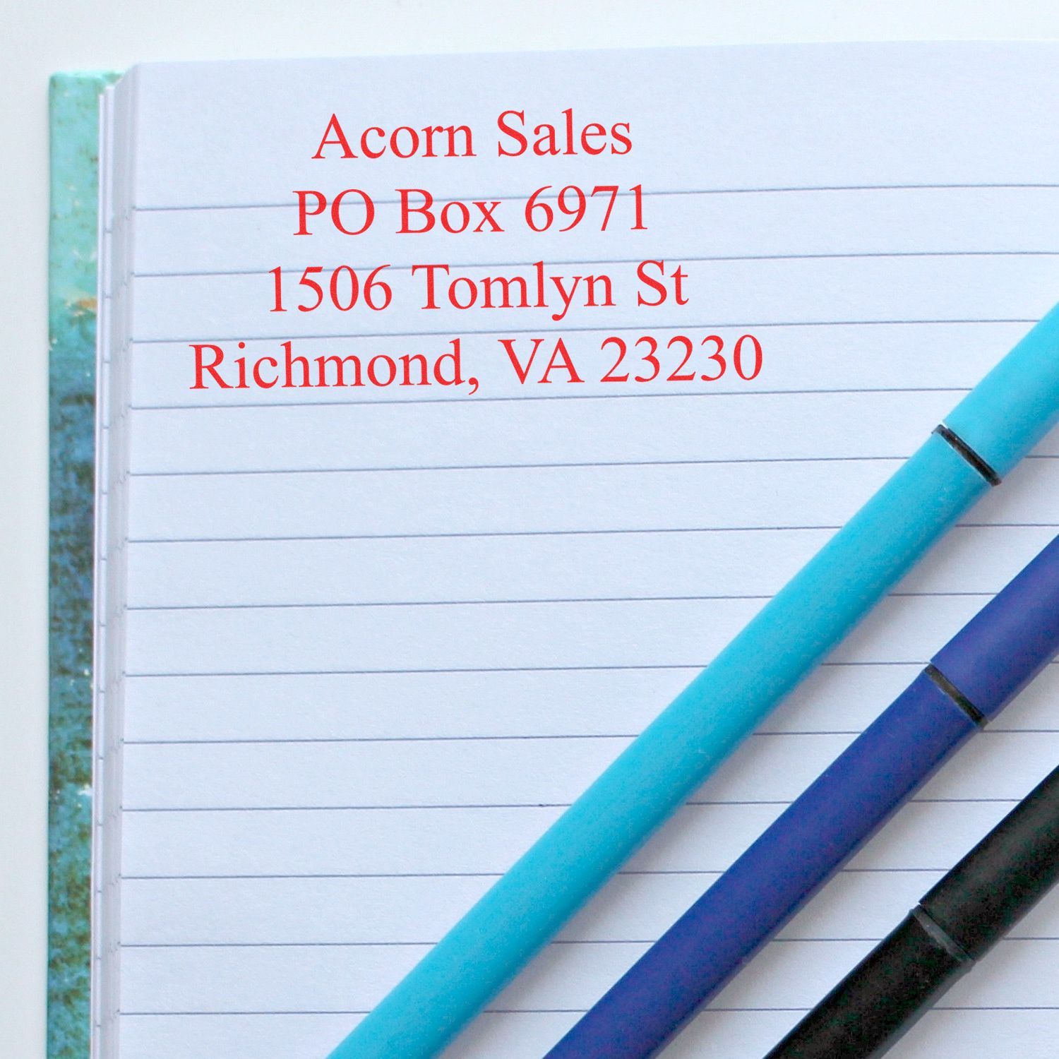 Close-up of lined notebook with Acorn Sales address in red text and colorful pens.