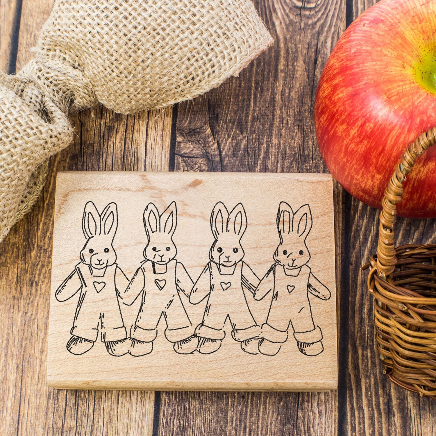 Rustic wooden bunny stamp with four rabbits illustration, apple, burlap, and wicker basket on a farmhouse wood table.