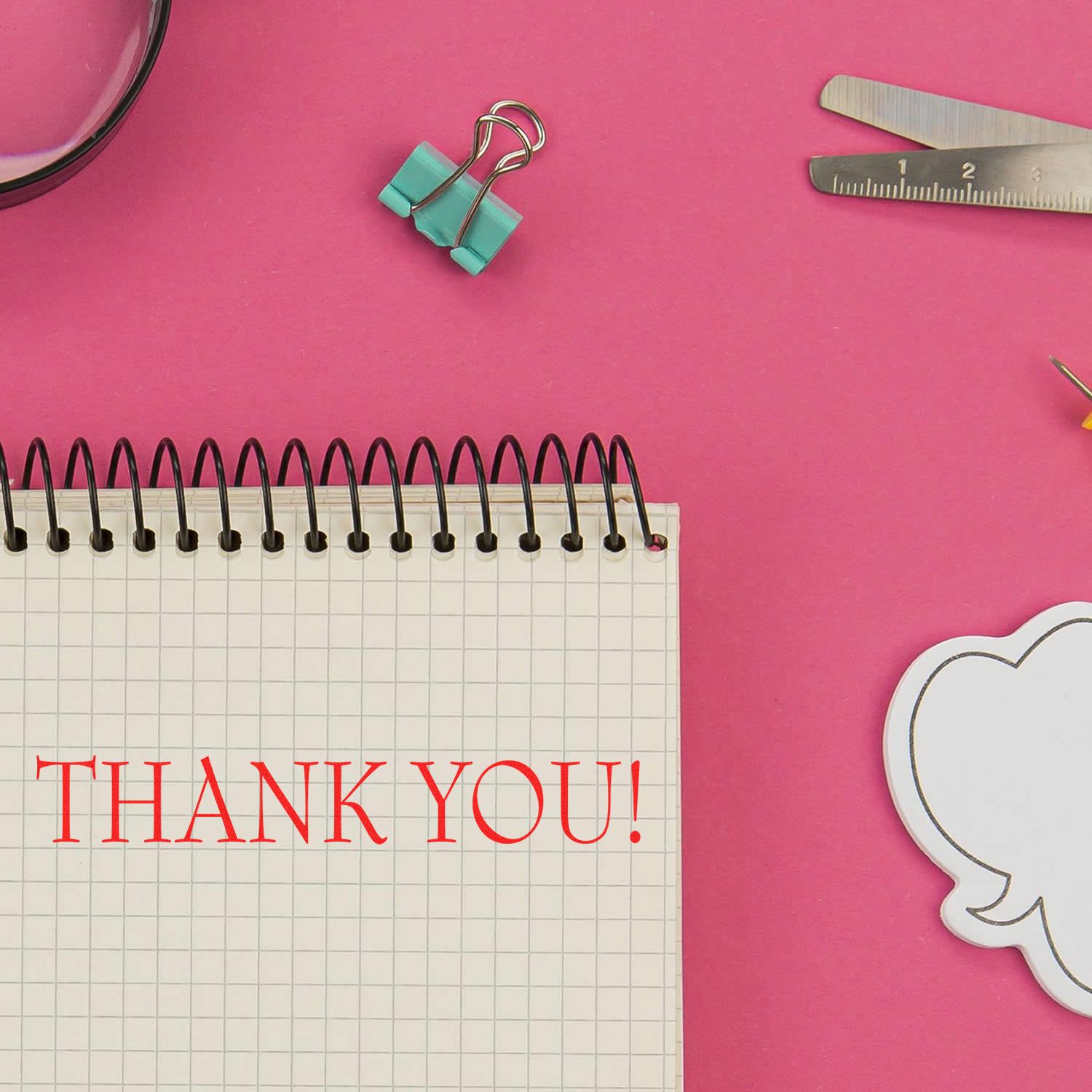 Close-up of a THANK YOU card message on grid notebook paper with binder clips, ruler, and pencil on pink desk, office stationery.