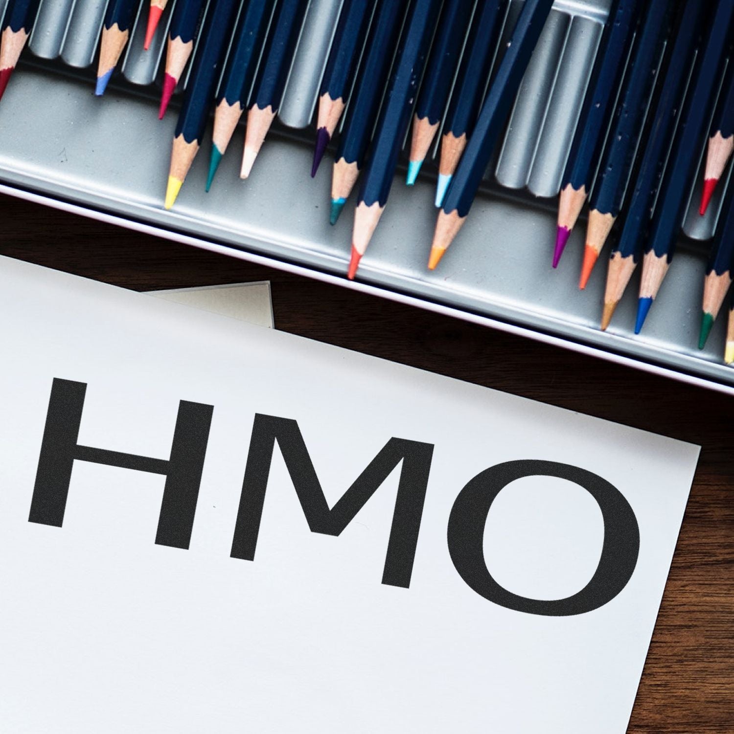 Colored pencils aligned above a sheet with bold HIMO lettering, representing writing, education, and creative design.