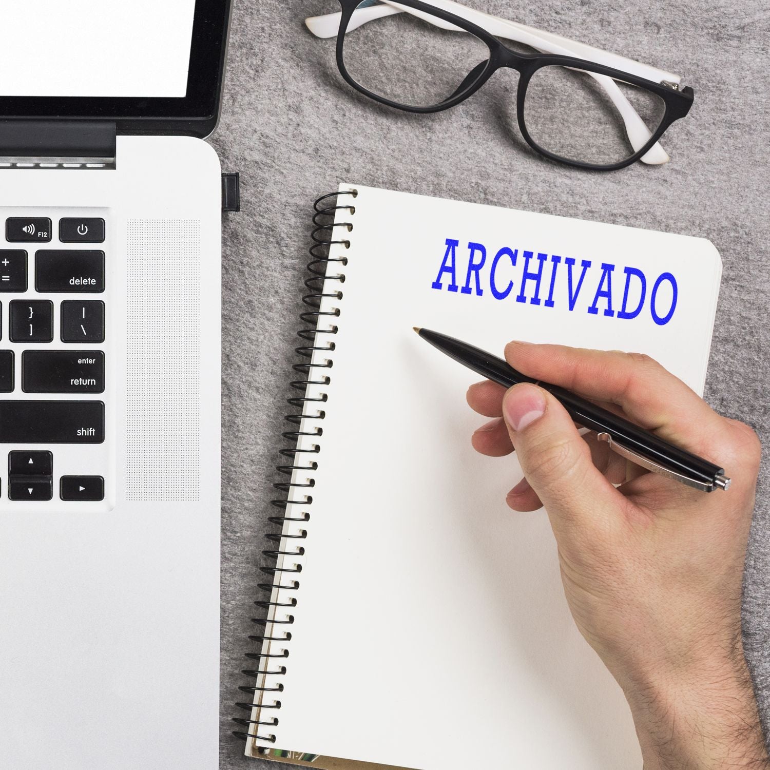 Hand writes ARCHIVADO in a notebook beside a laptop, highlighting document archiving and record keeping for office organization.