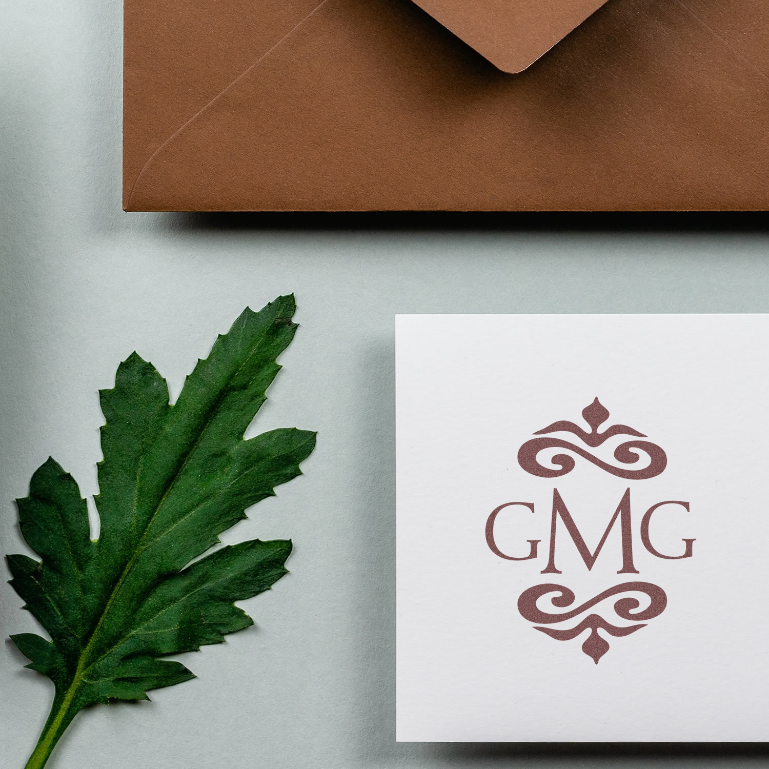 Custom monogram rubber stamp impression with ornate GMG design on stationery, beside a brown envelope and green leaf.