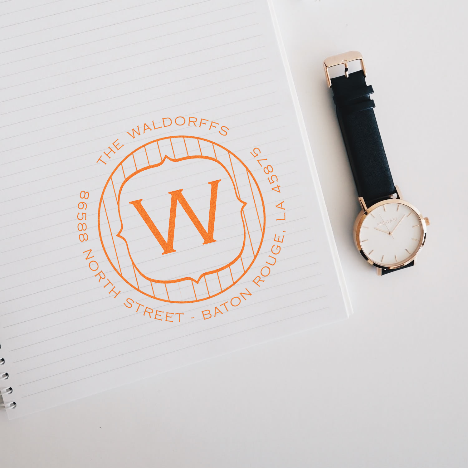 Custom monogram rubber stamp with family name and address stamped on lined notebook beside a wristwatch.