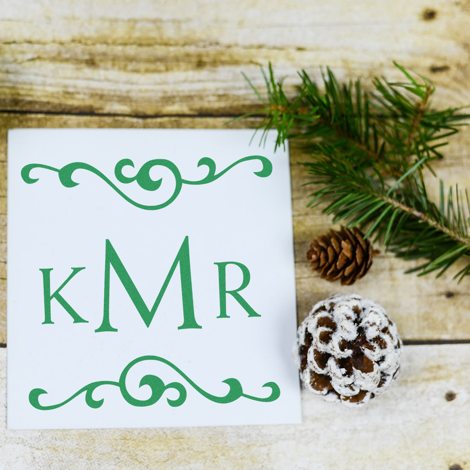 Custom KMR monogram rubber stamp with decorative flourish, displayed on white card with pinecone and evergreen on rustic wood.
