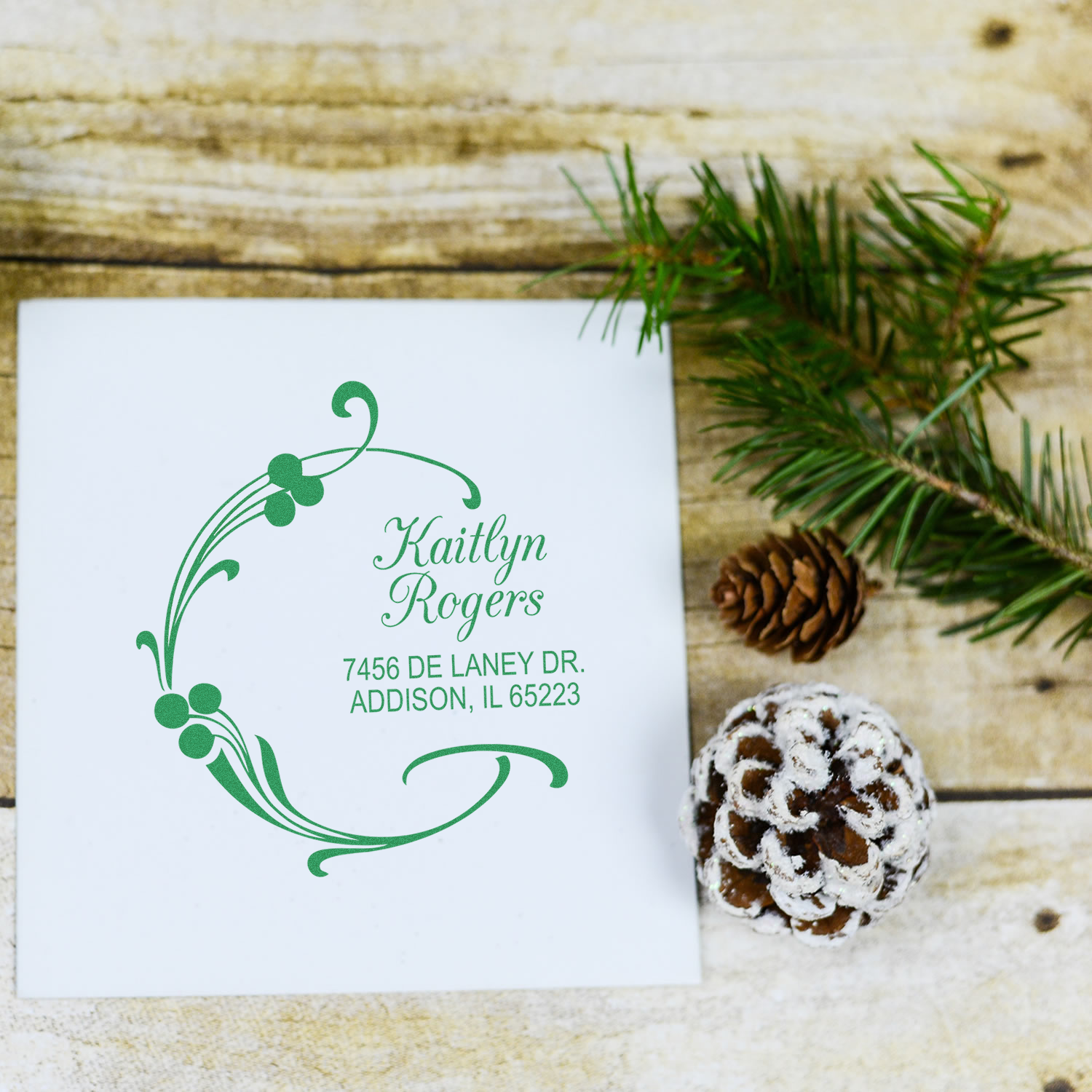 Custom holiday return address rubber stamp with green floral wreath design, personalized name and address, styled with pine branch and cone.
