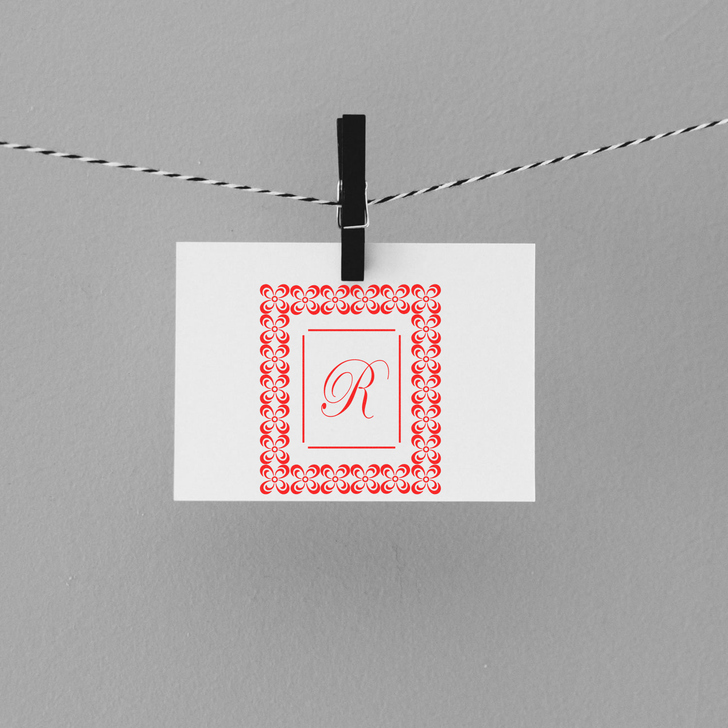Clothespin pinned card with ornate red border and monogram letter R, decorative greeting invitation for weddings or events on string.