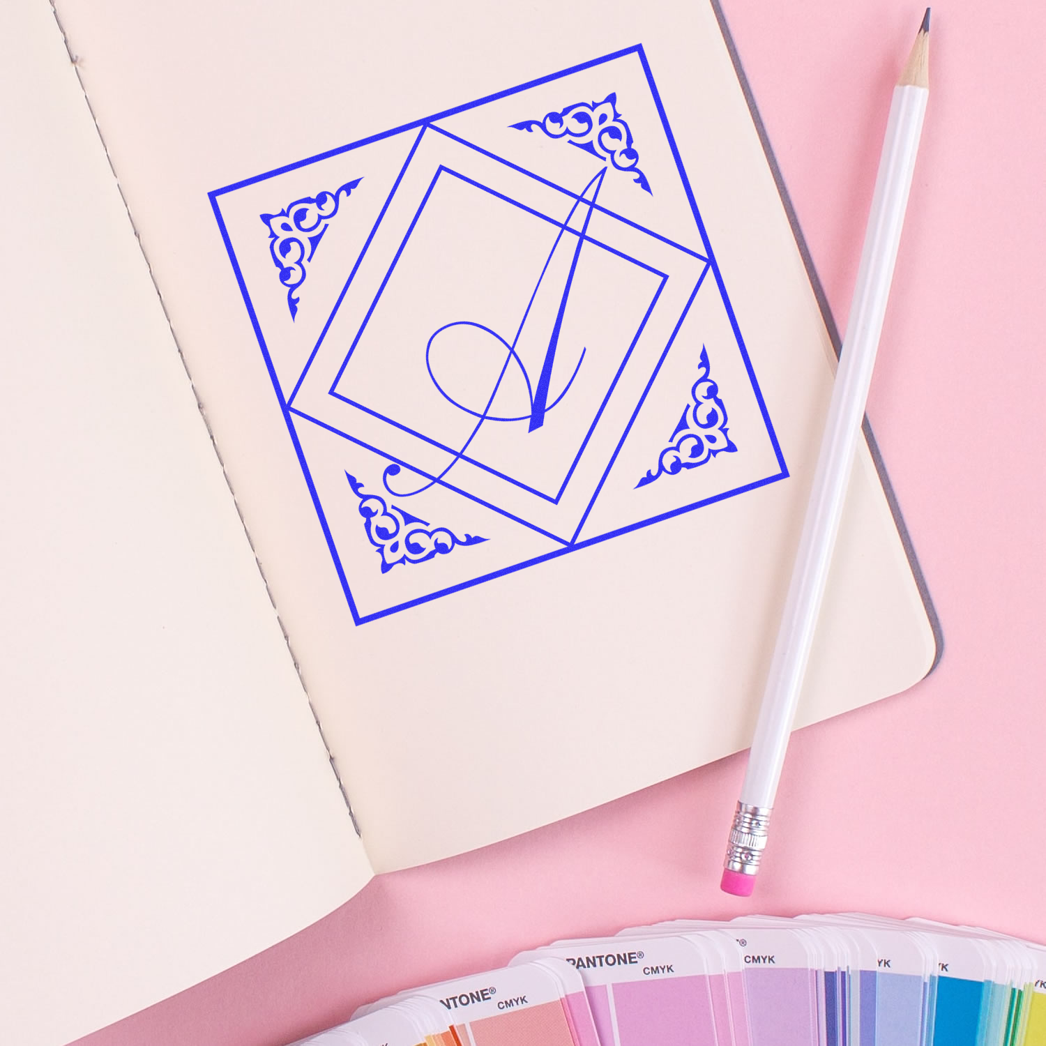 Custom rubber stamp monogram design in blue ink on notebook, with pencil and color swatches for stationery branding.
