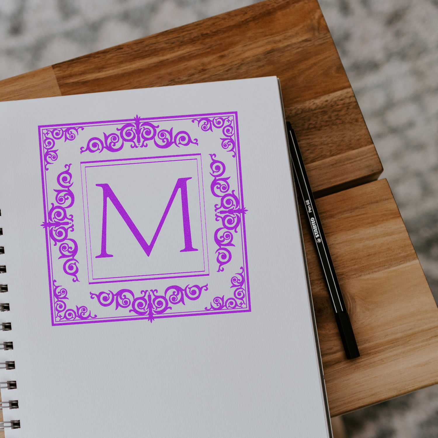 Custom decorative monogram rubber stamp with letter M in purple on a sketchbook, ideal for personalized stationery and branding.