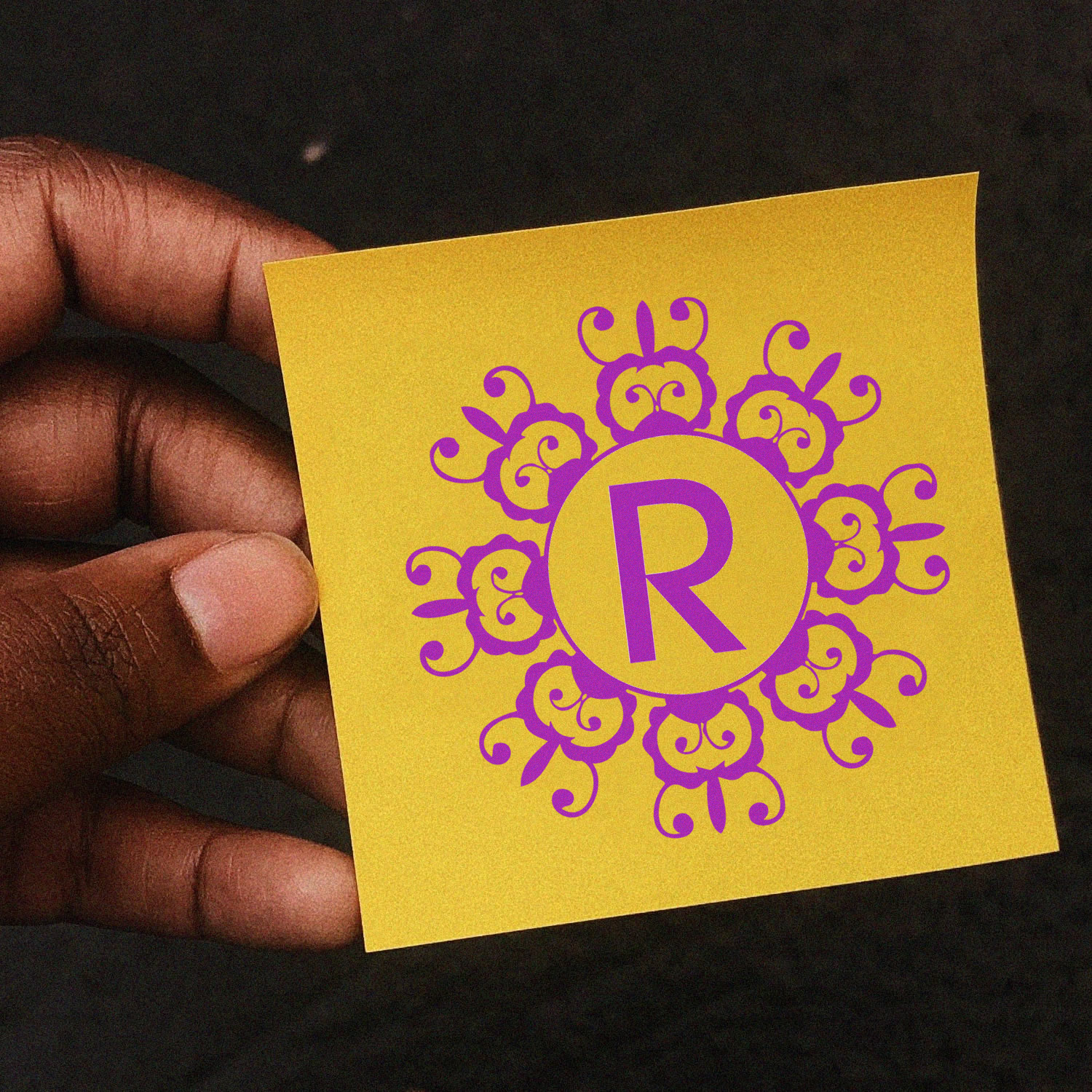 Hand holding yellow paper with purple ornate monogram R, showing a custom decorative rubber stamp design.