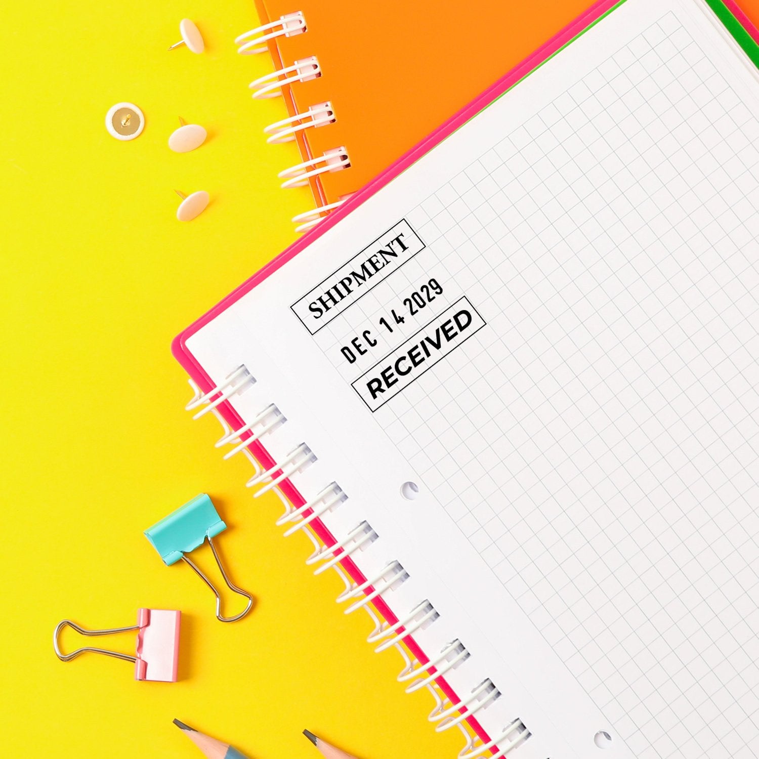 Colorful spiral notebook with Shipment Received stamp on bright yellow background, surrounded by stationery and binder clips.