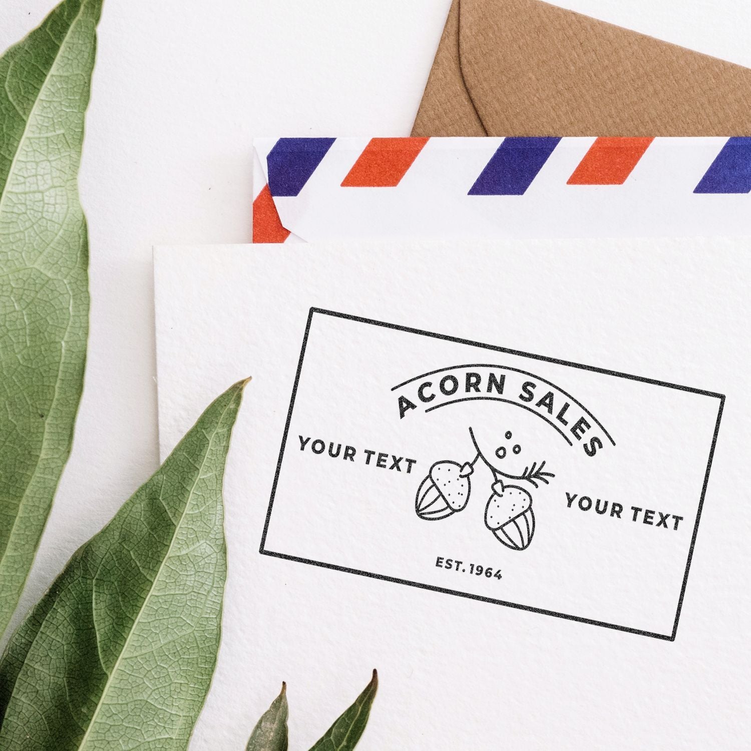 Custom acorn logo stamp card on white paper with kraft envelope and green leaves, for personalized branding and product labels mockup