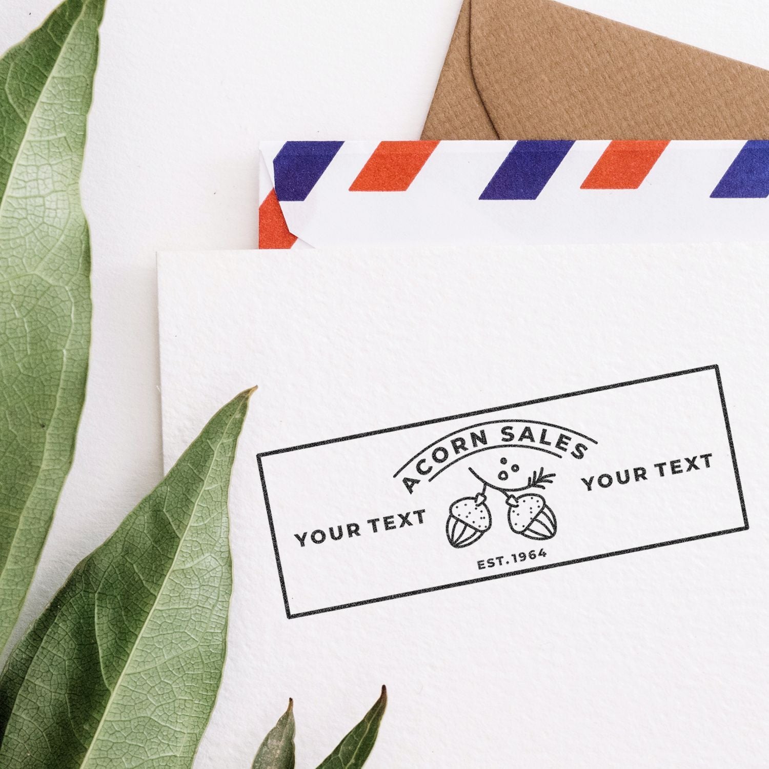 Custom stamp-style product label mockup on white paper and kraft envelopes with green leaves, Acorn Sales branding.