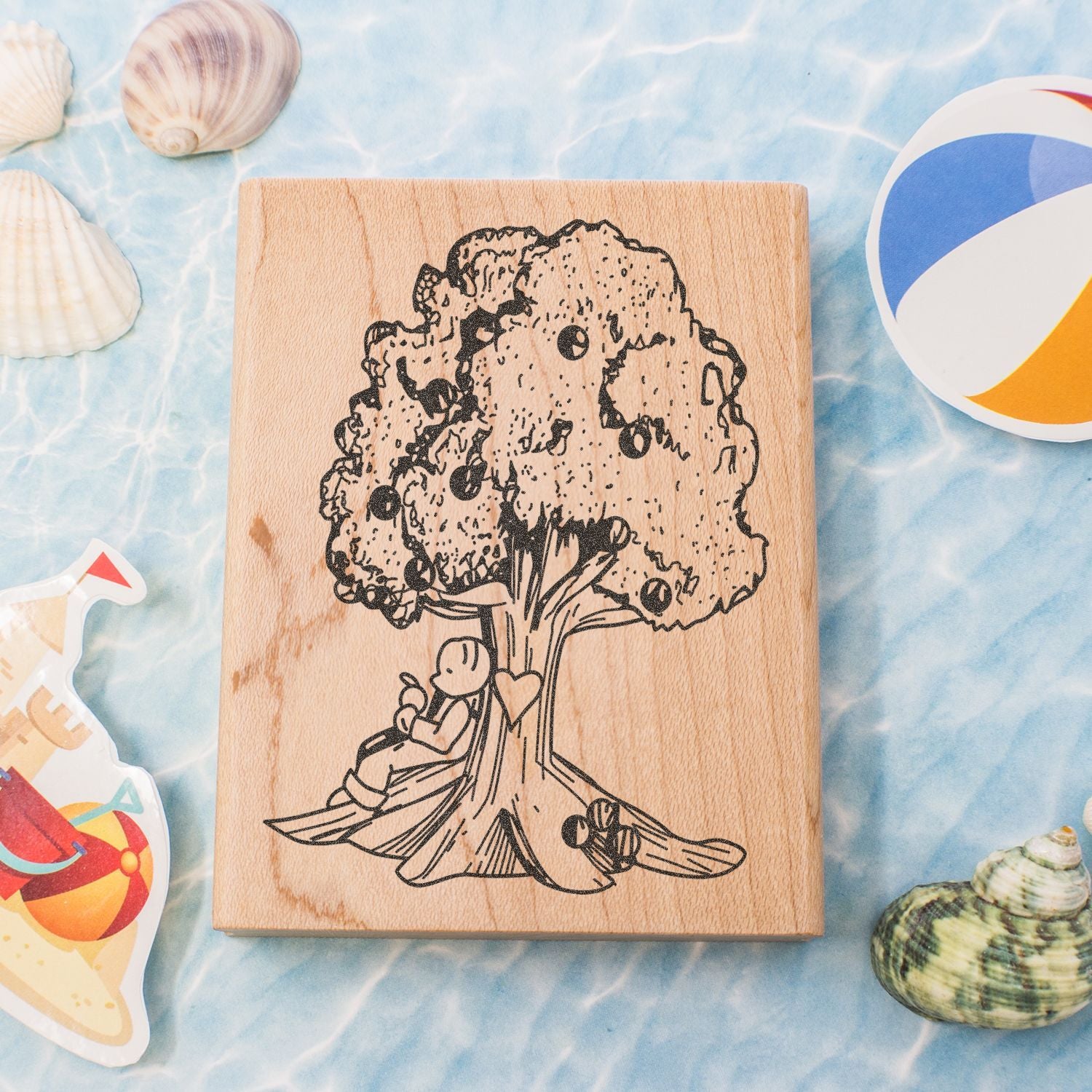 Wooden rubber stamp with a tree and child illustration, styled with seashells and beach toys on a blue summer background.