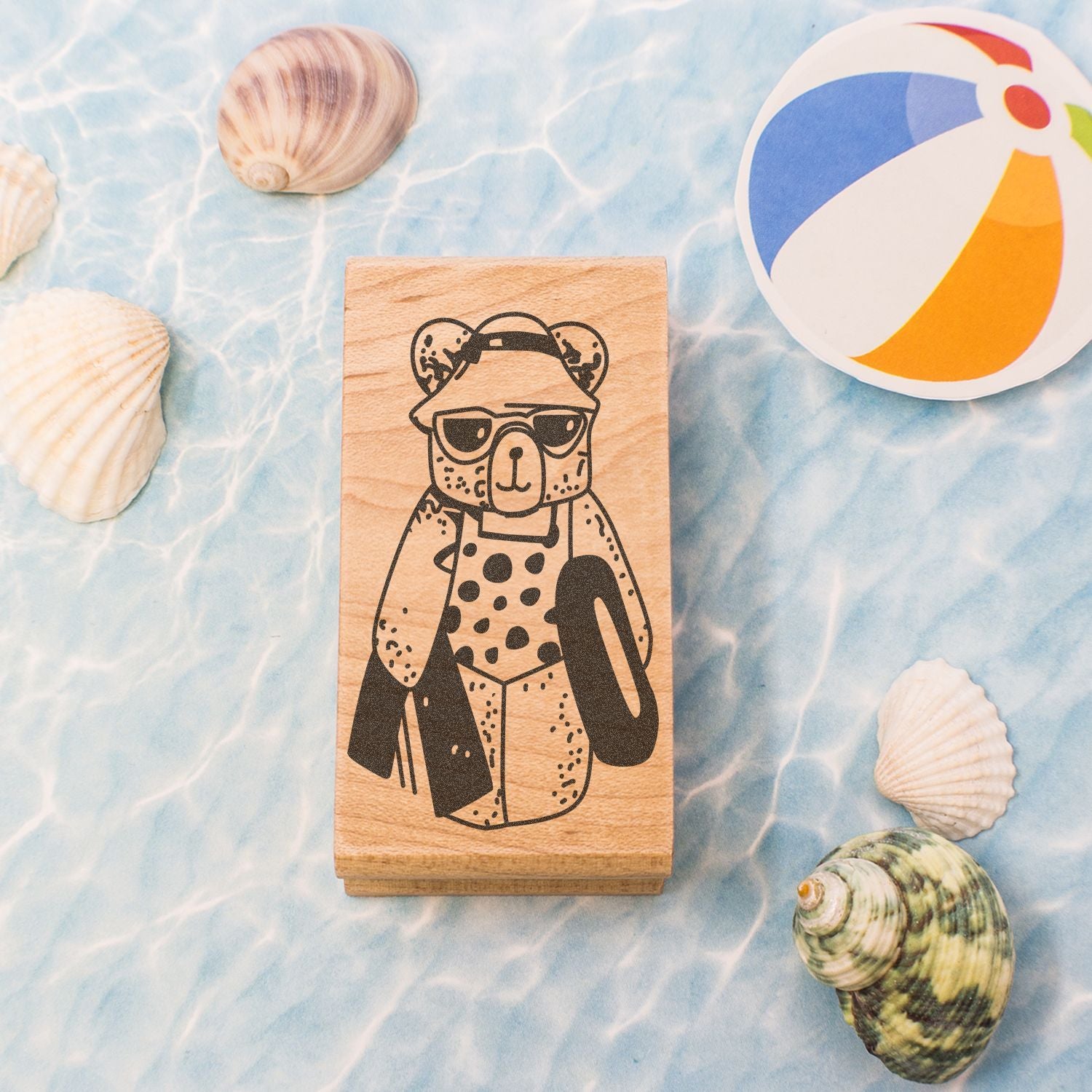Wooden bear stamp with sunglasses and hat on a beach-themed background with seashells and a colorful beach ball.