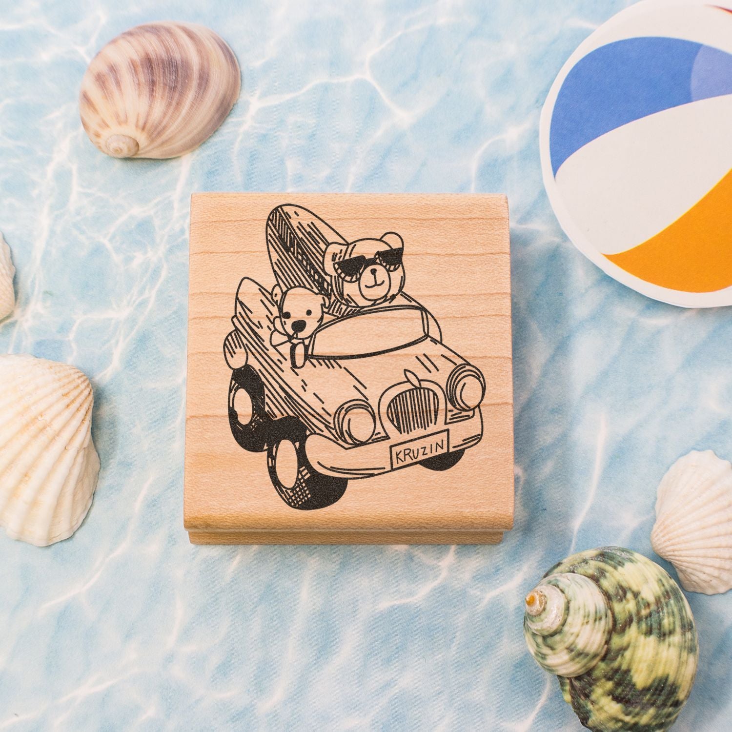 Wooden beach-themed stamp with teddy bear driving a car, surrounded by seashells and a beach ball on a blue water background.