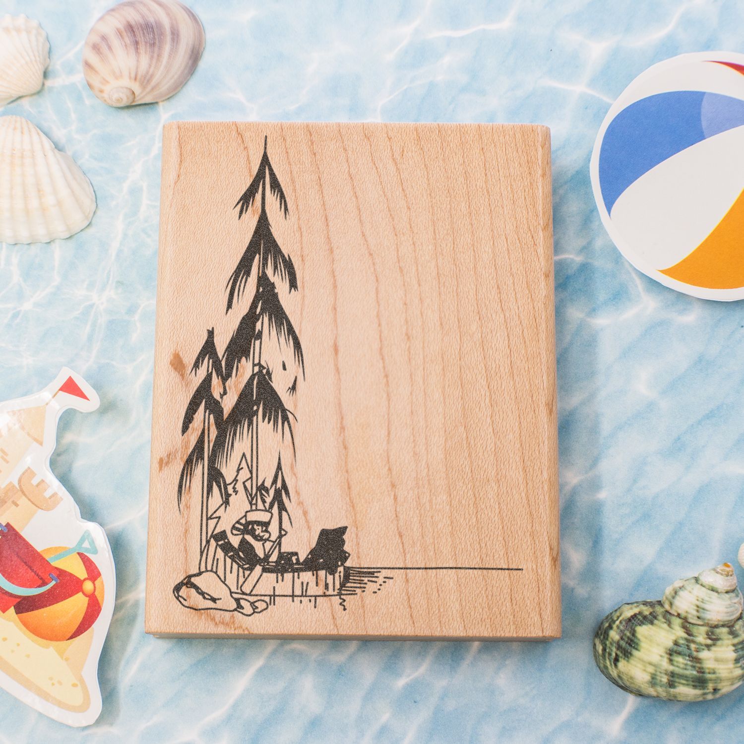 Wooden engraved serving board with pine tree design, surrounded by seashells and beach-themed decor on a light blue water background.