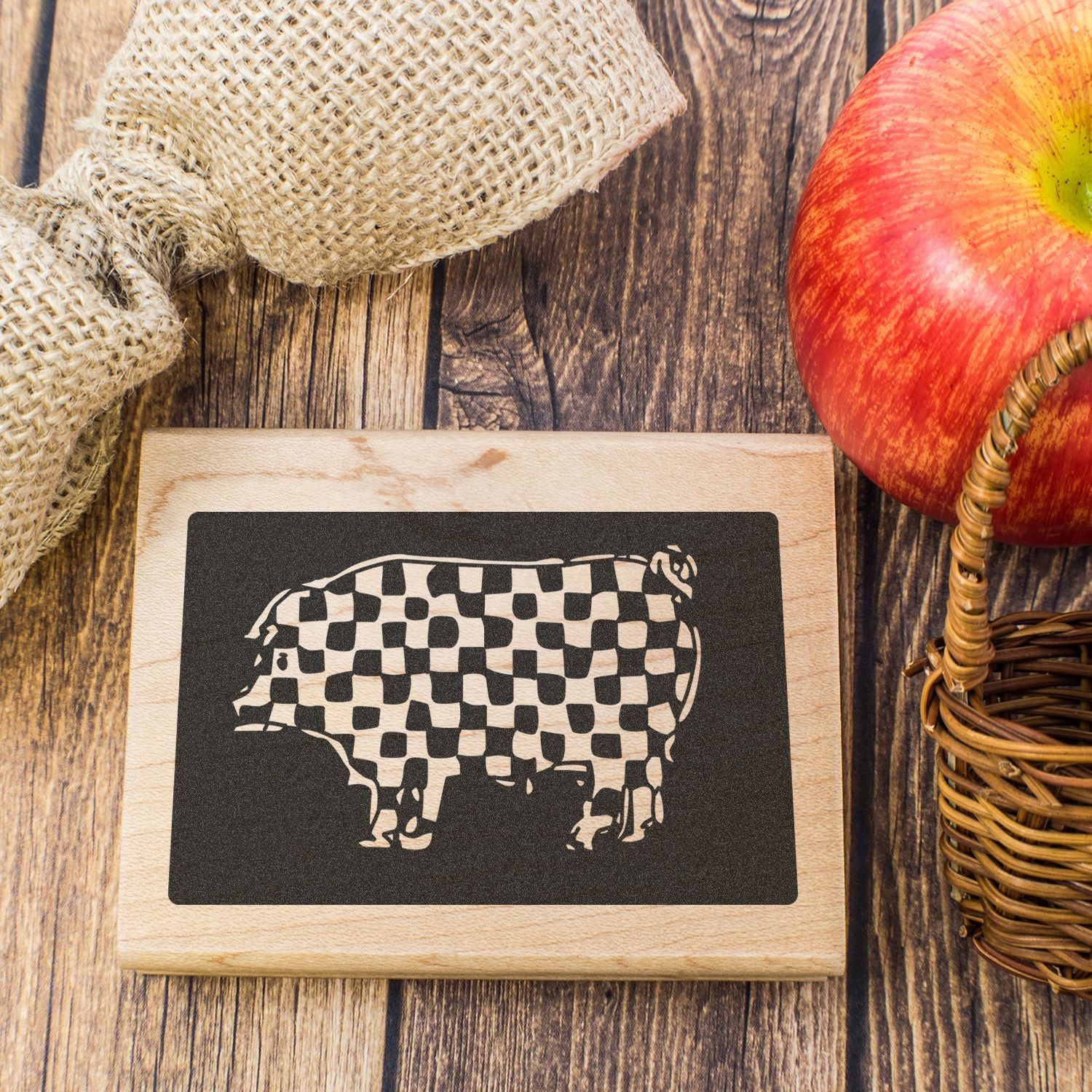 Rustic farmhouse wooden sign with checkered pig cutout, apple, burlap, and wicker basket on a wood table.