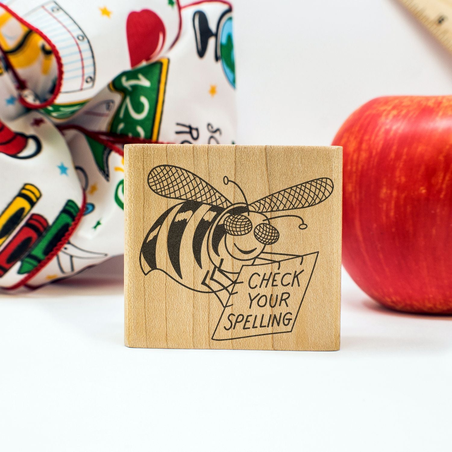 Wooden Check Your Spelling stamp with bee illustration, school supplies, and red apple on a white background.