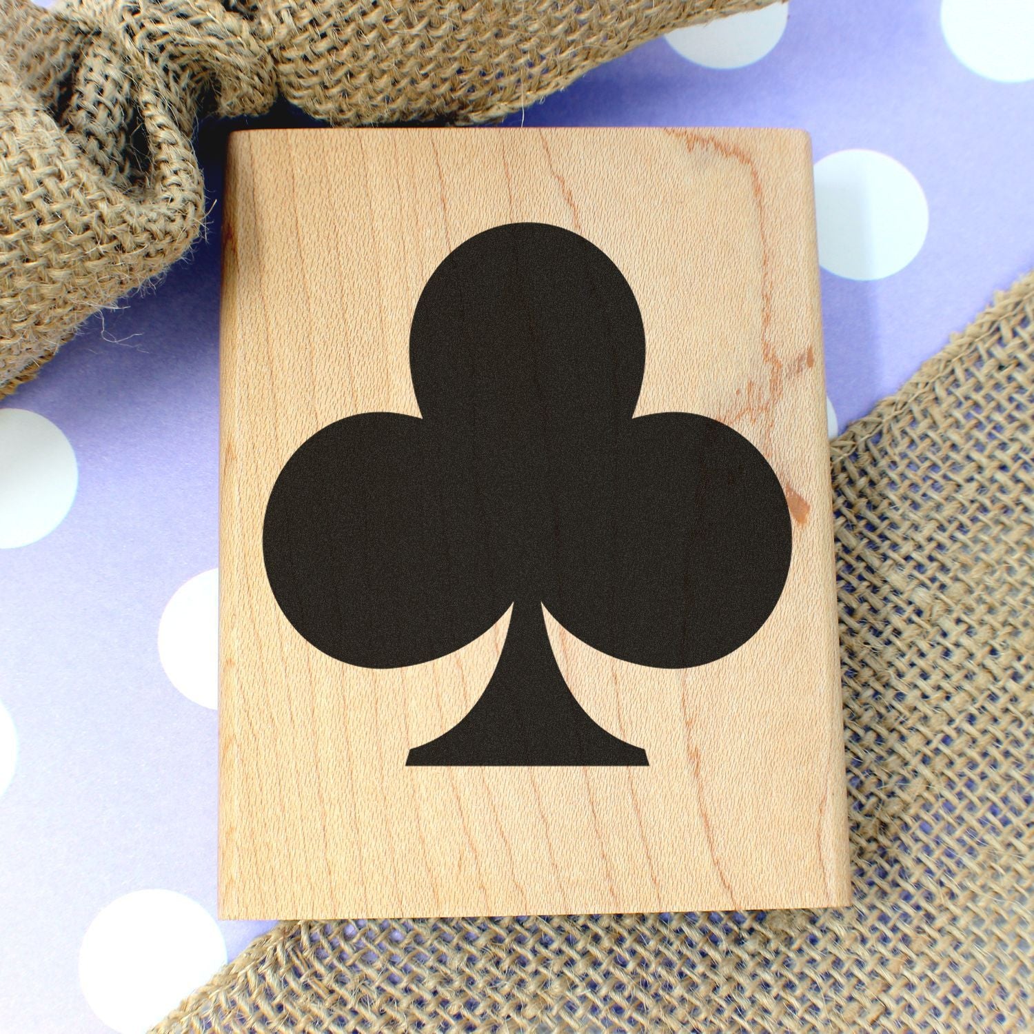 Wooden playing card with black clubs symbol on a purple polka dot background with burlap texture.