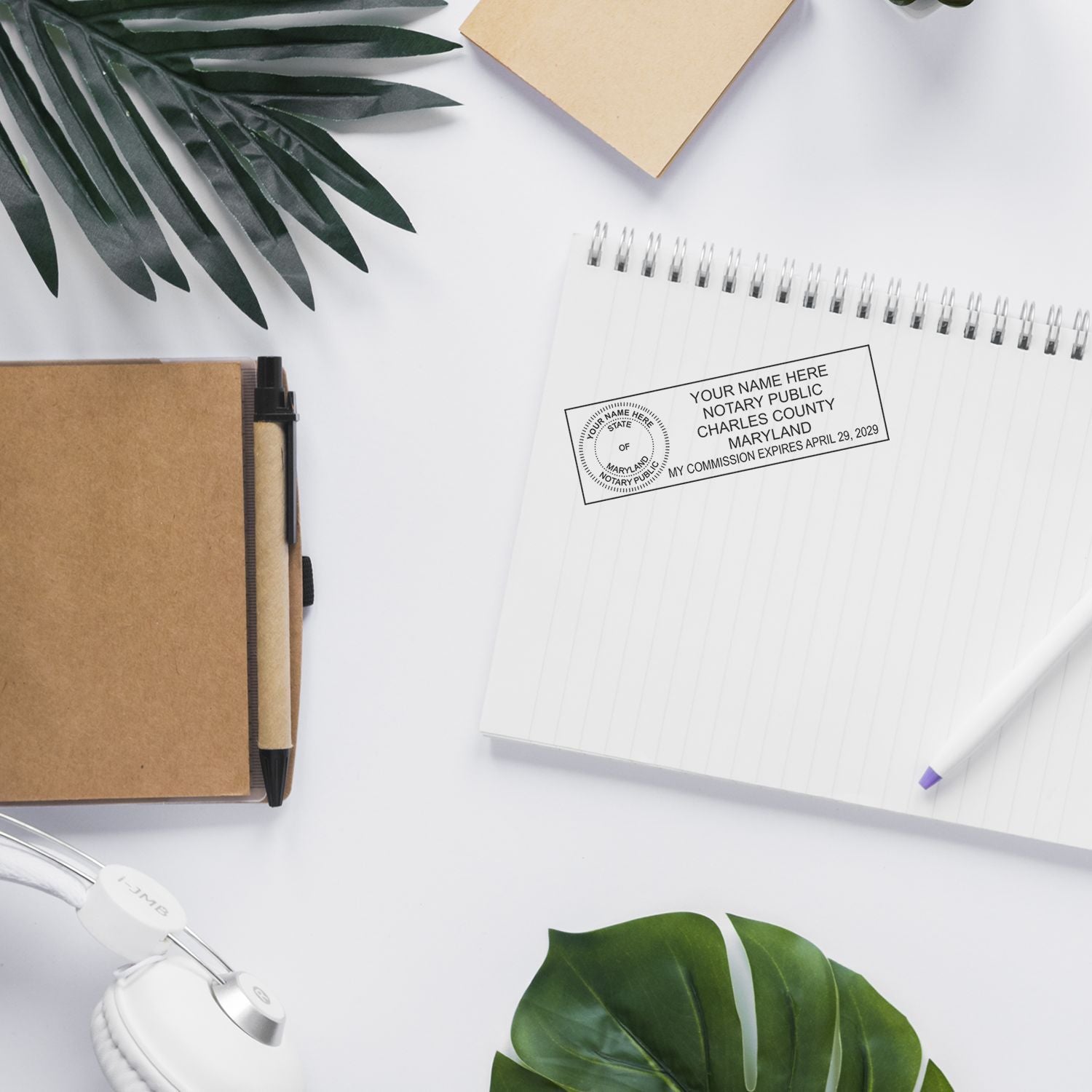Flat lay of a Maryland notary public stamp on a spiral notebook with office stationery, pen, headphones, and tropical leaves on a white desk.