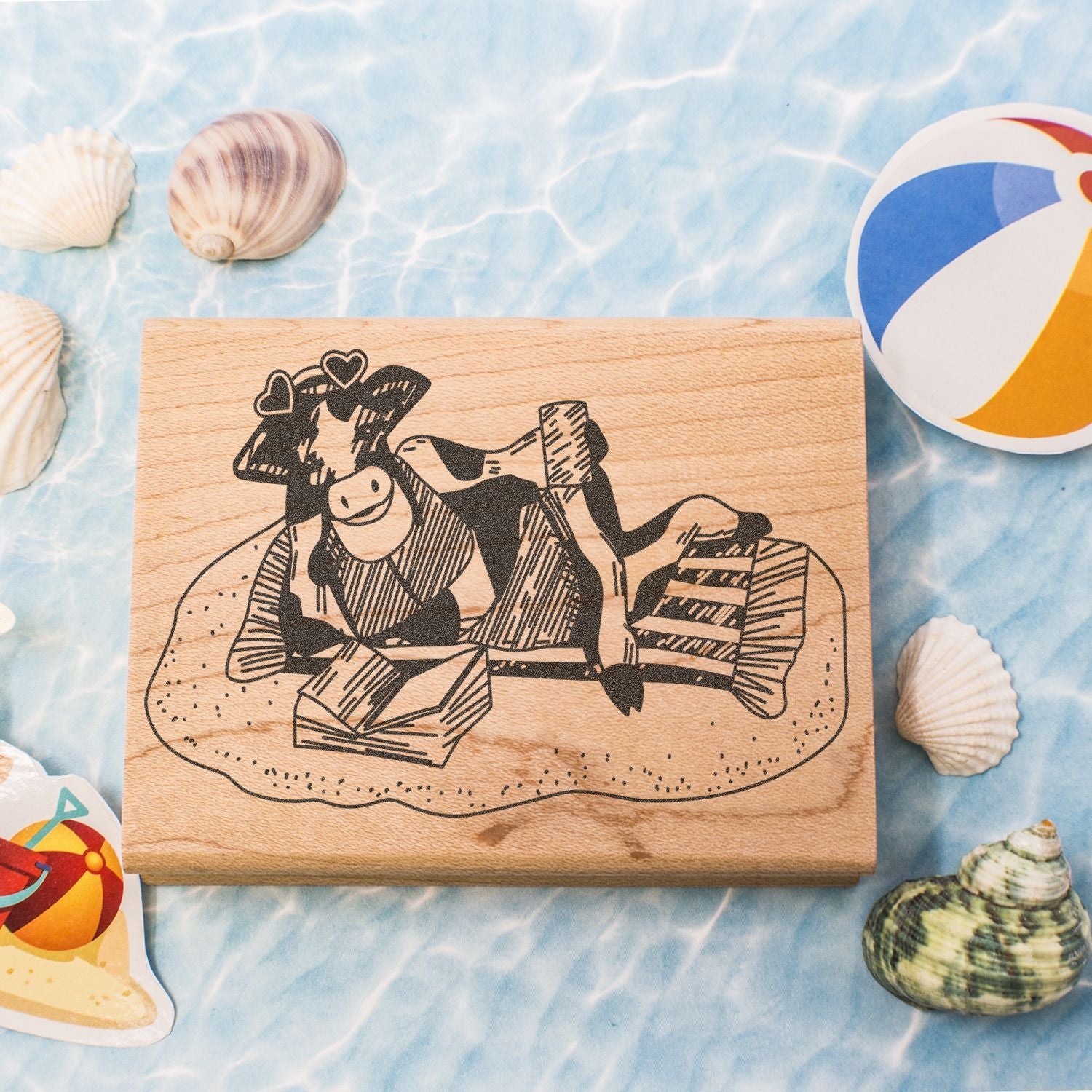 Wooden rubber stamp with whimsical cow beach design, surrounded by seashells and a colorful beach ball on a summer ocean background.