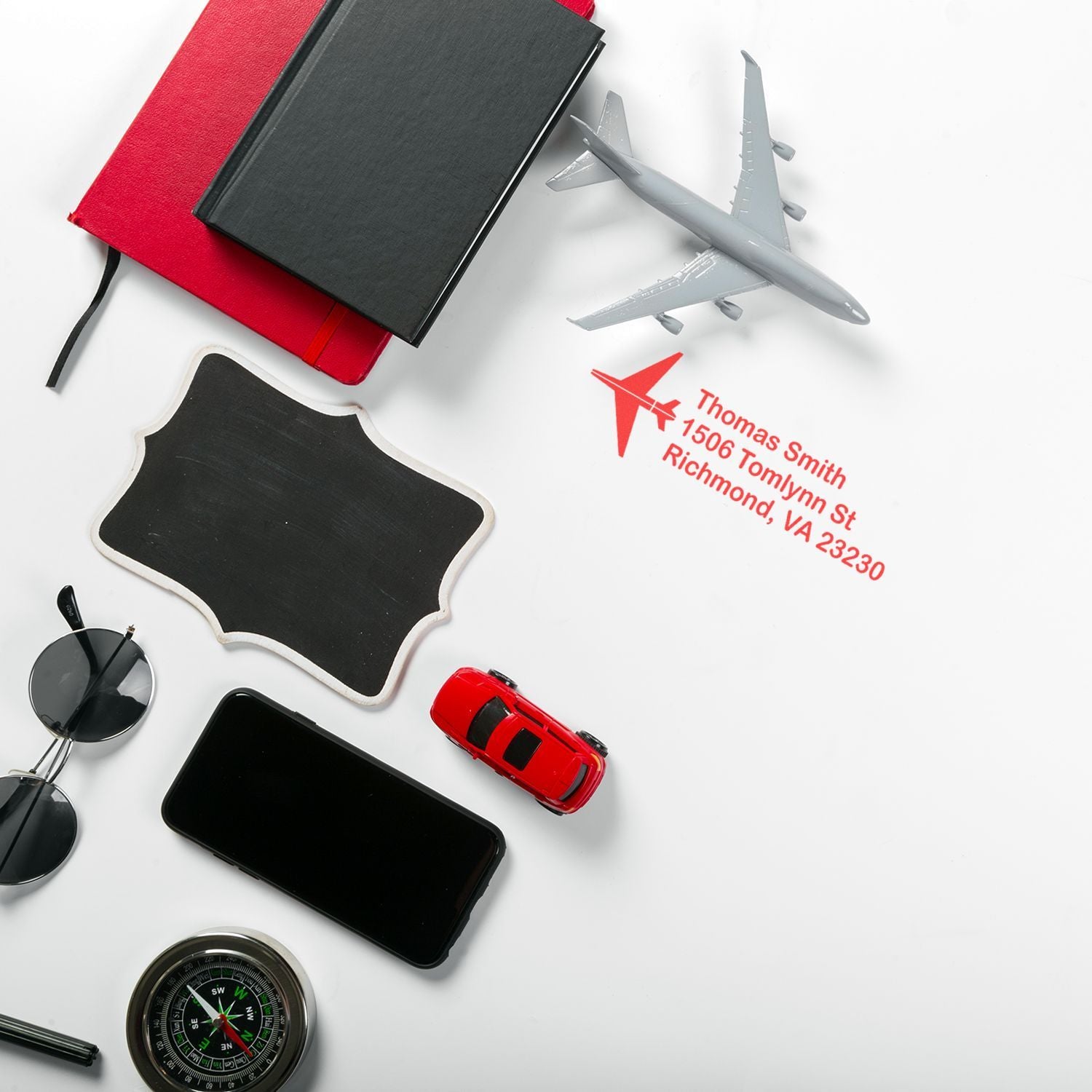 Travel flat lay featuring a custom address rubber stamp with airplane icon, notebooks, plane model, phone, compass and sunglasses.
