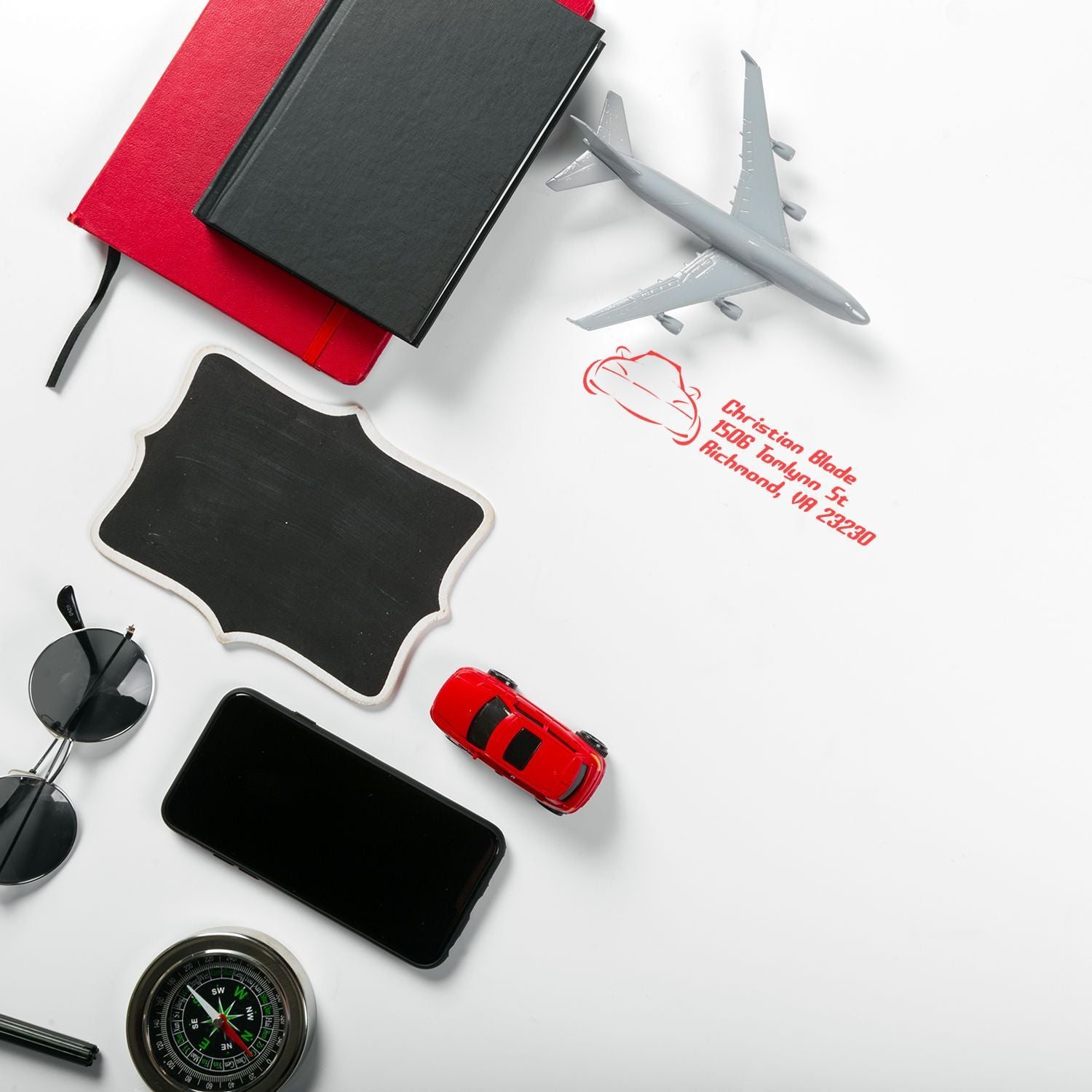Personalized rubber address stamp imprint on a travel-themed flat lay with notebooks, airplane, compass, phone and sunglasses.