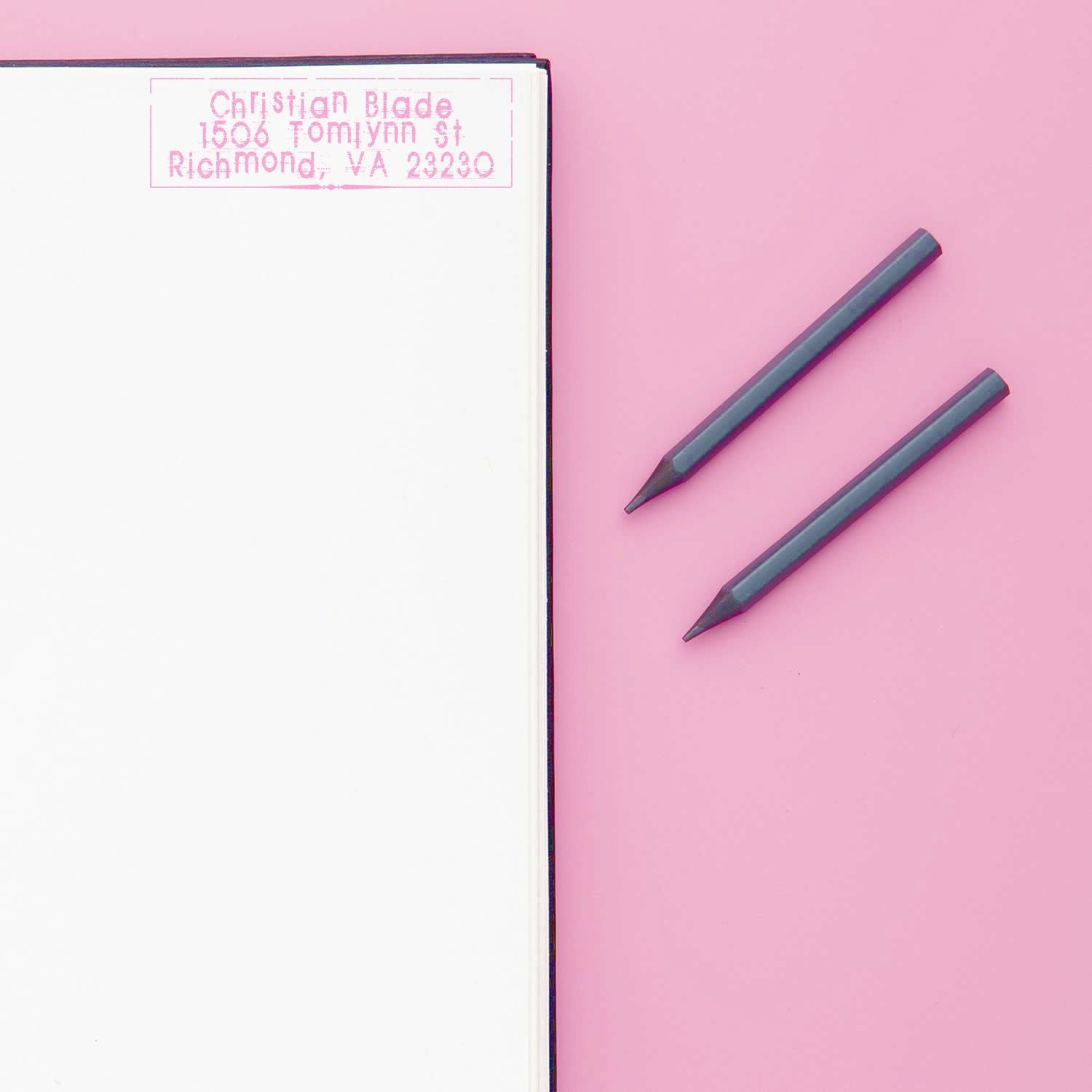 Custom address rubber stamp impression on notebook page, with two stamping tools on a pink background.