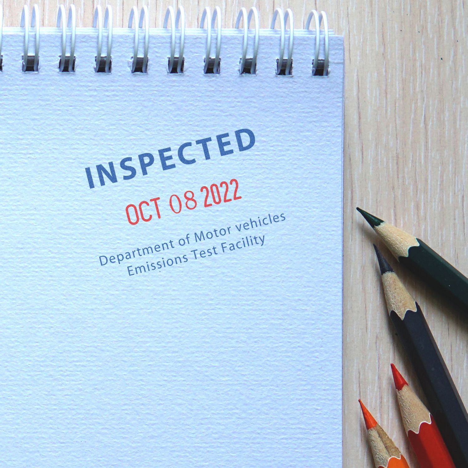 Spiral notebook stamped Inspected with vehicle emissions test date and colored pencils on a wooden desk.