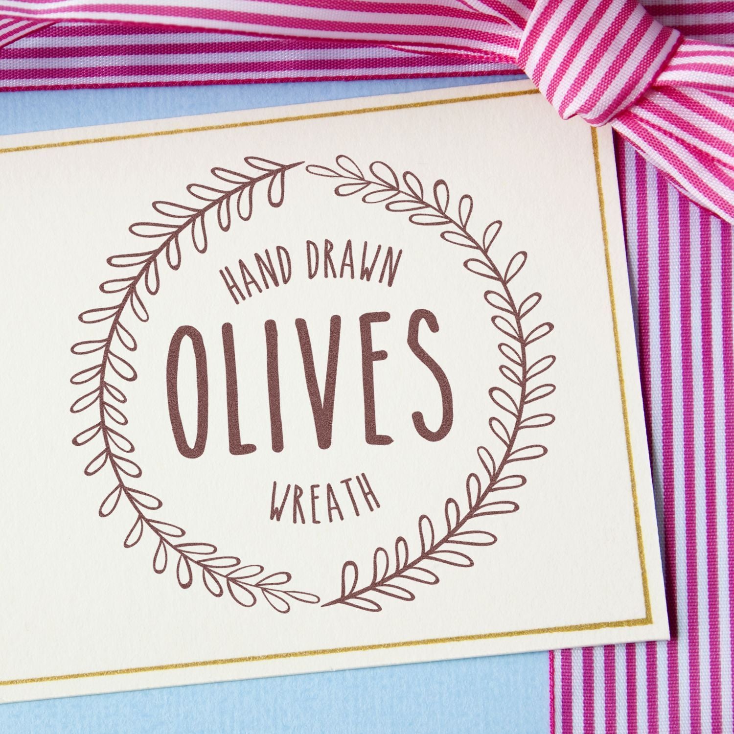 Hand-drawn olives logo lettering on greeting card with olive branch wreath, illustrated olive print on pink striped fabric.
