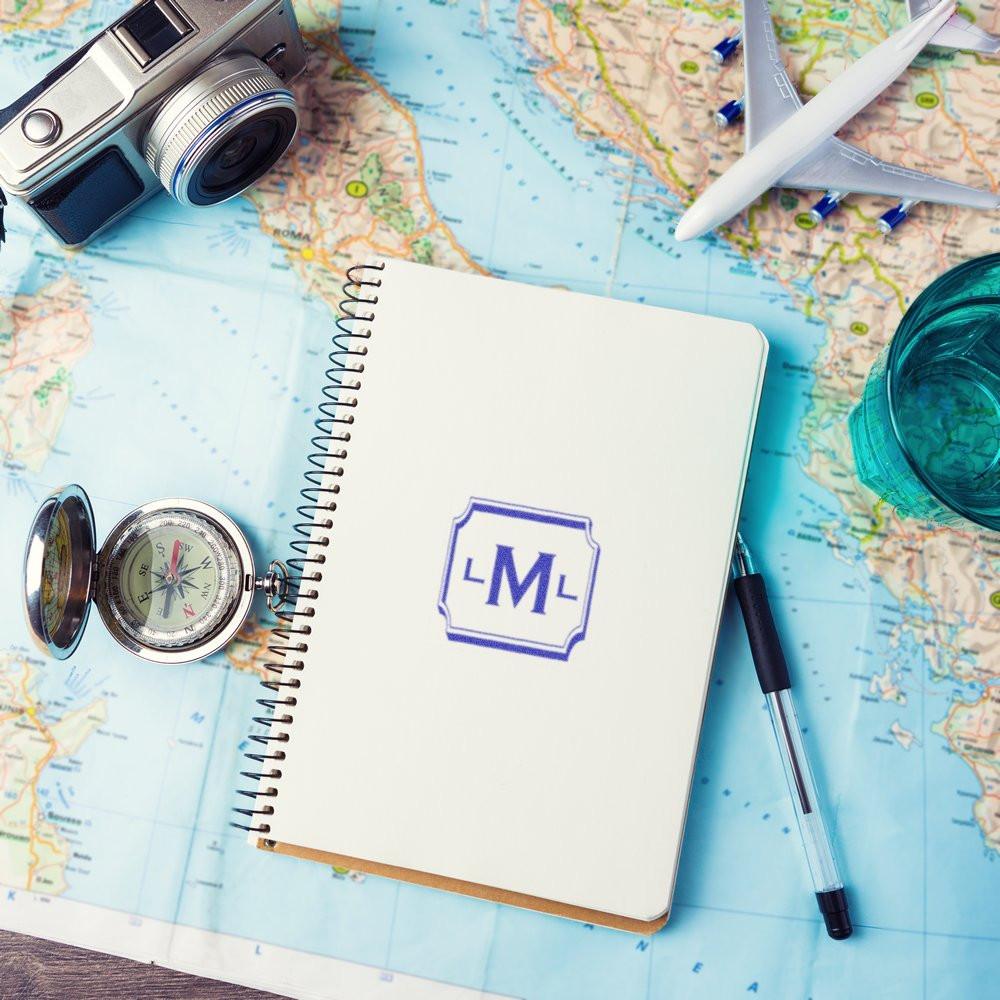Custom monogram rubber stamp on a travel notebook, with map, compass, camera and plane for personalized travel branding.