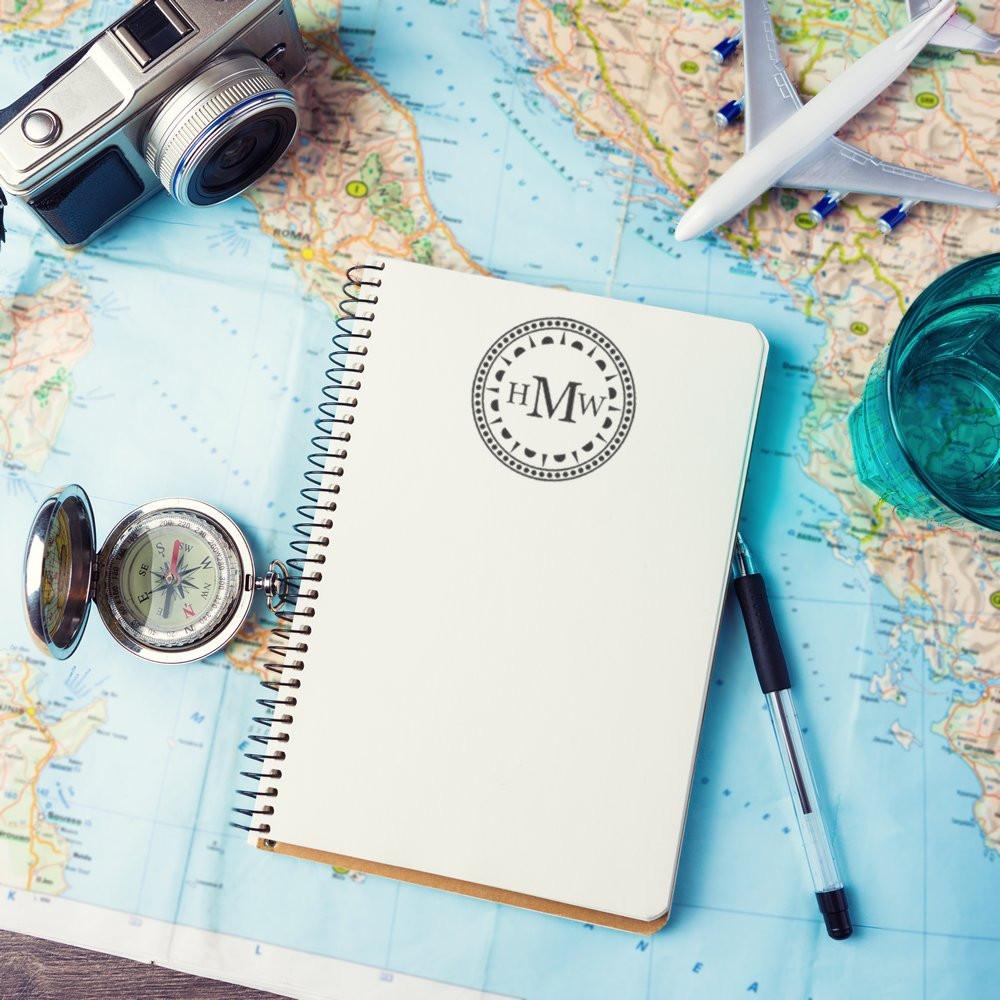 Personalized rubber stamp on blank travel journal, surrounded by map, camera, compass and airplane for trip planning.