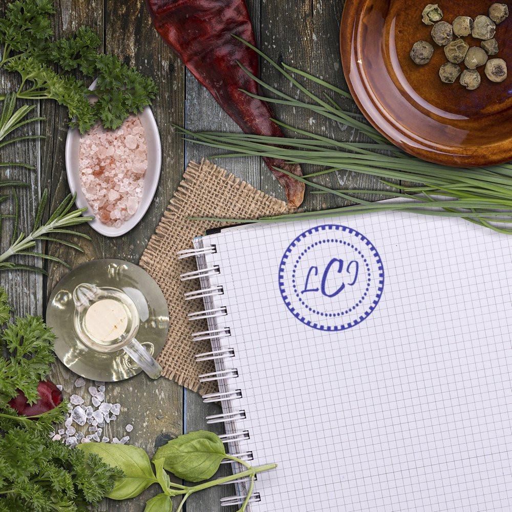 Circular monogram rubber stamp on graph paper notebook, styled with herbs and spices for branding and packaging.
