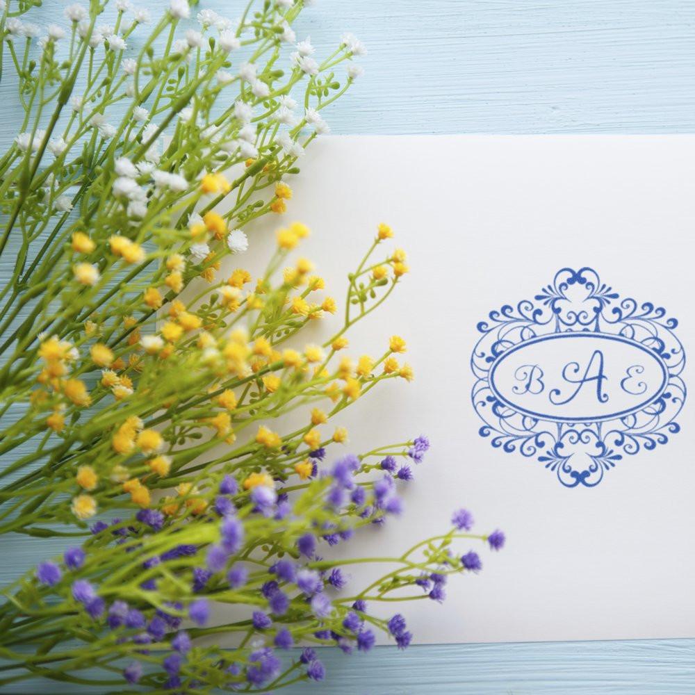 Personalized floral monogram rubber stamp with ornate blue initials on a white card beside pastel flowers.