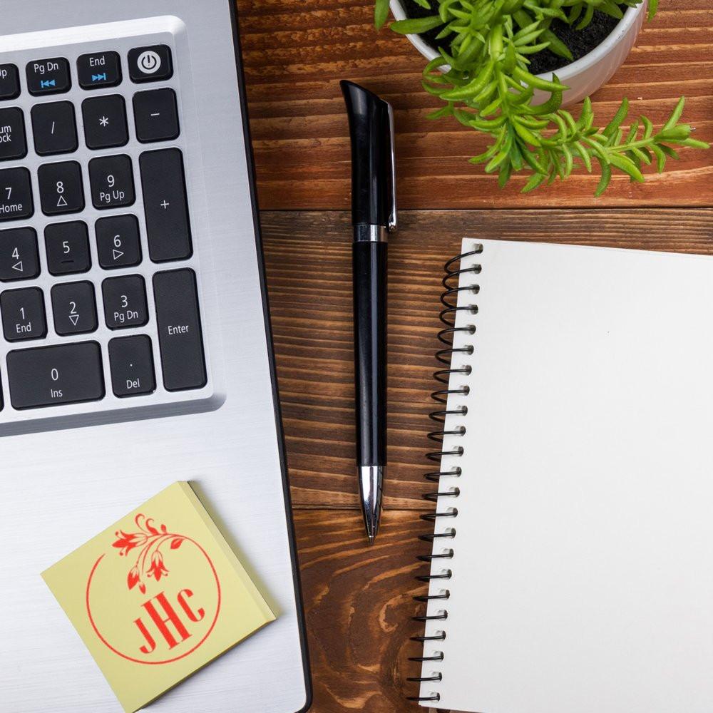 Custom monogram rubber stamp on sticky note beside laptop, notebook, and pen for personalized office branding.