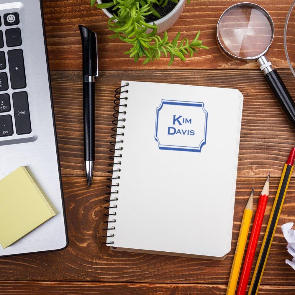 Personalized rubber stamp imprint on notebook with Kim Davis name, shown on a desk with laptop, pen, and stationery.