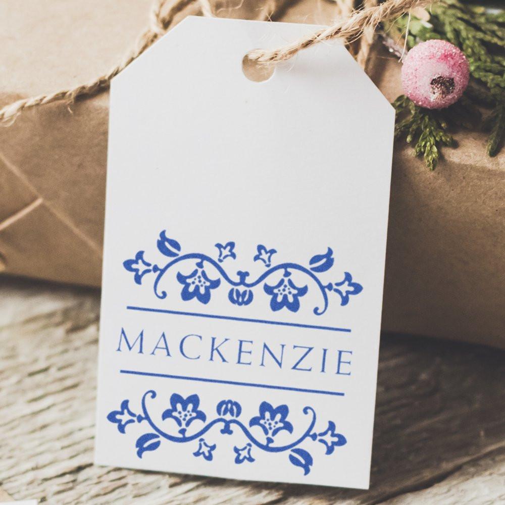 Custom rubber stamp on white gift tag with blue floral monogram and Mackenzie name for personalized packaging.