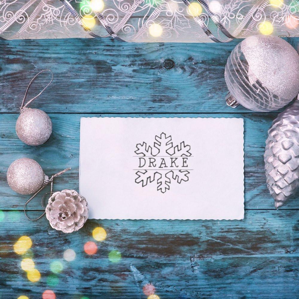 Winter holiday greeting card with snowflake logo reading DRAXE, surrounded by Christmas ornaments and pine cone on blue wood.
