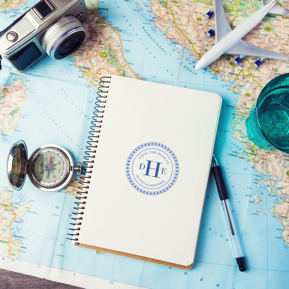 Custom rubber stamp on a travel notebook over a world map, with camera, compass, plane, and pen for personalized branding.