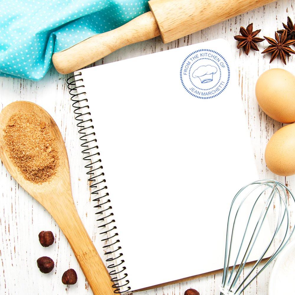 Custom chef rubber stamp on recipe notebook with eggs, whisk, spoon, and spices for baking branding.