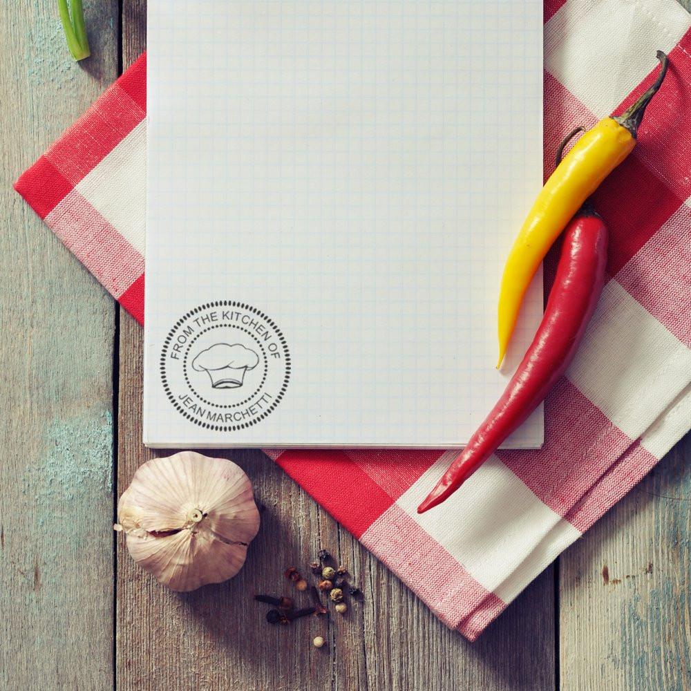 Personalized rubber stamp on recipe notebook with chef hat logo for kitchen branding, surrounded by chili peppers and garlic.