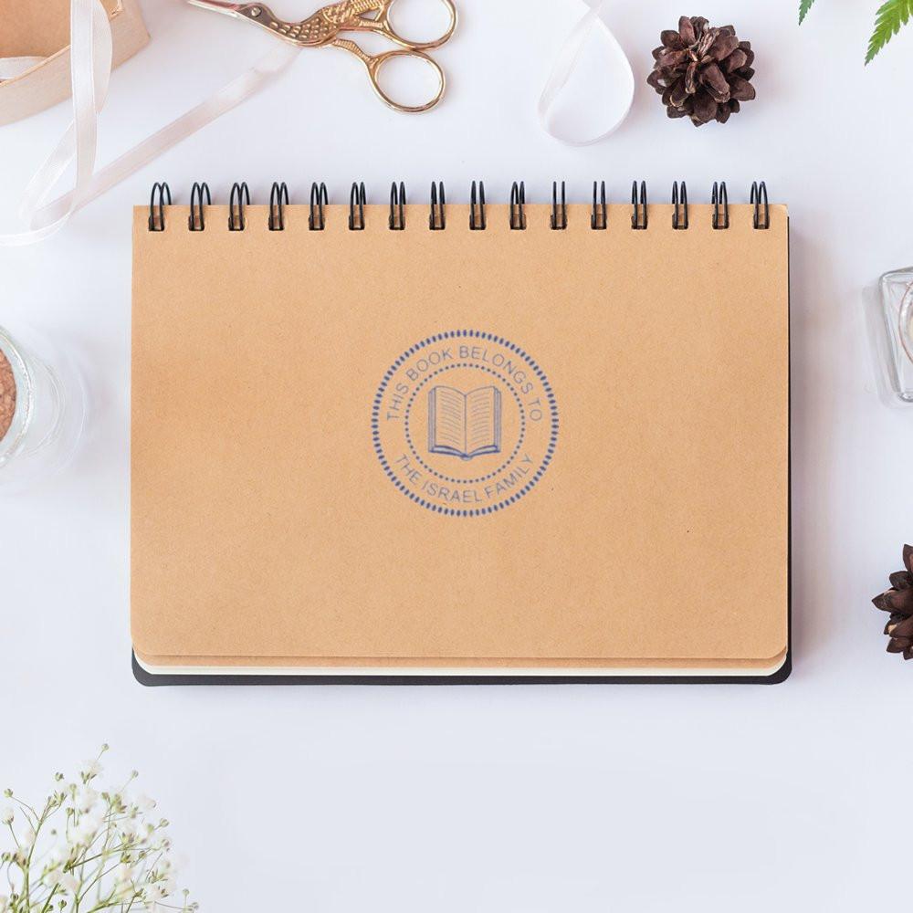 Custom rubber stamp impression on notebook cover with a This Book Belongs To book ownership design for libraries and readers.