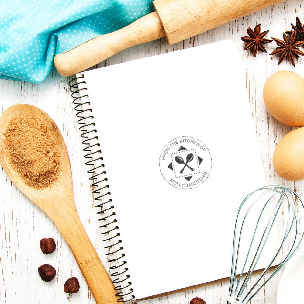Personalized kitchen rubber stamp on a recipe notebook, styled with baking tools for custom baker branding and packaging.