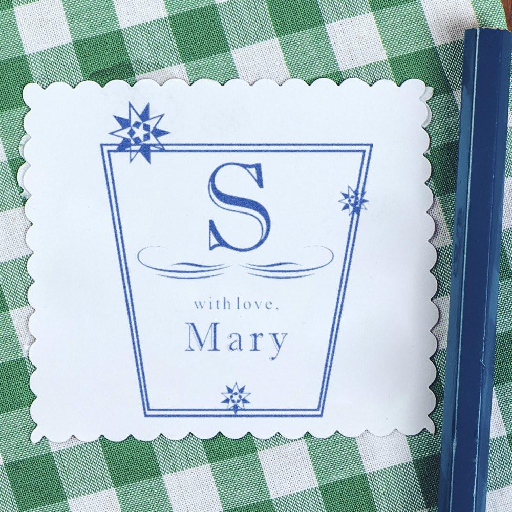 Personalized blue letter S on white scalloped stamp with with love, Mary, set on green gingham fabric background.