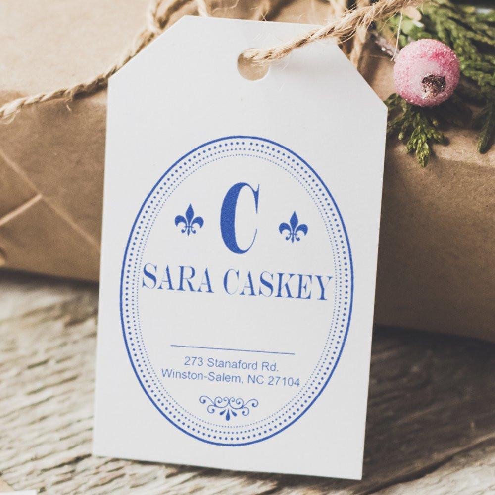 Custom monogram address rubber stamp on white gift tag, featuring blue oval name design for personalized packaging.