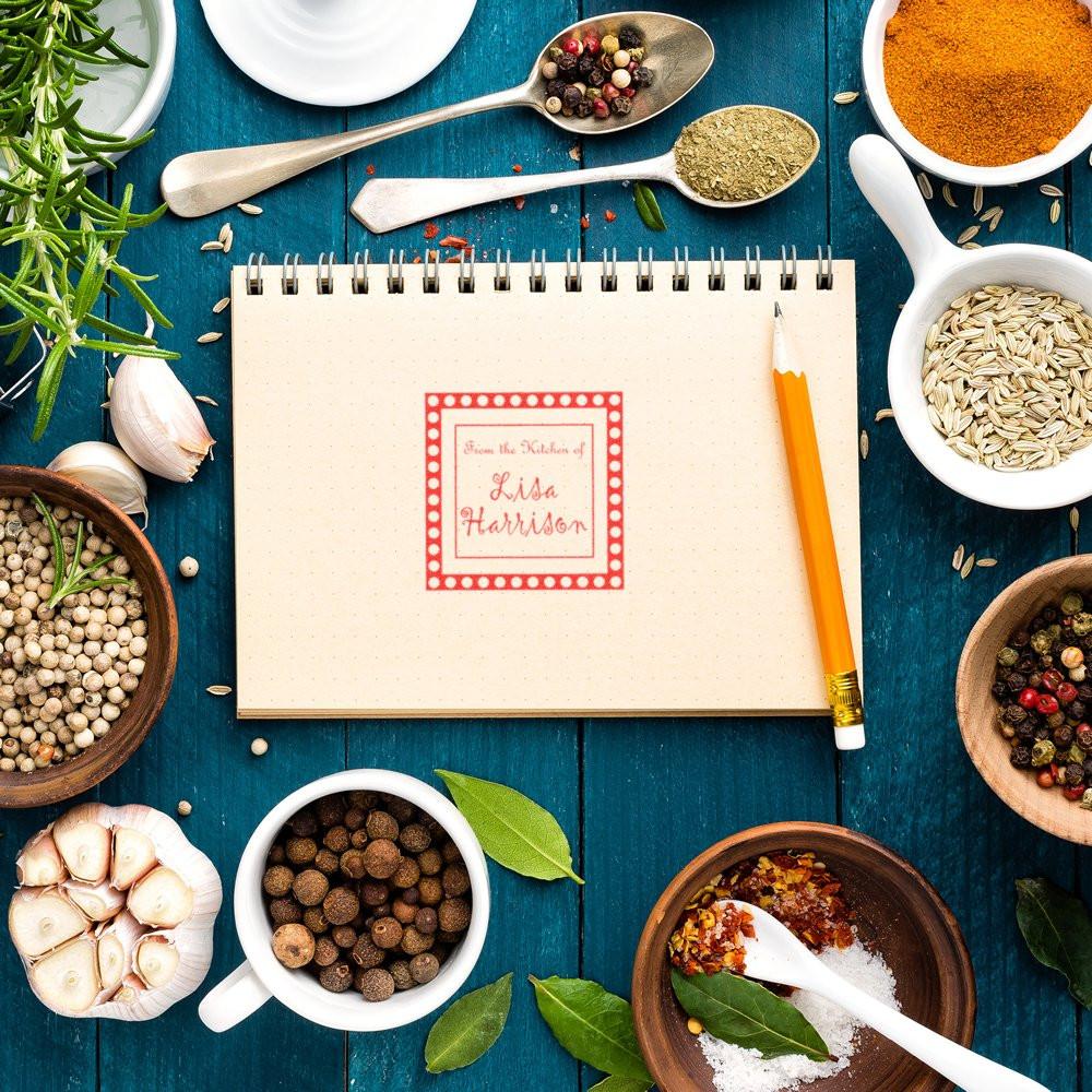 Personalized kitchen rubber stamp on a recipe notebook surrounded by spices and herbs.